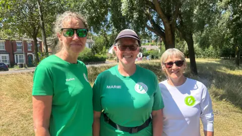 Three women, two in green t-shirts and one in a white t-shirt, are pictured outside in front of a grassy bank with houses beyond the grassy section. 