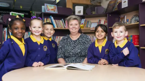 St Mary's CE Primary School A woman is sat at a table, with a book in front of her, and three children to her left and two to her right. The woman has light-coloured hair flicked to one side. The children wear blue and yellow uniforms