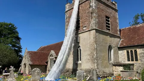 A picture of an ancient church. A cascade of white created to look like a waterfall is coming from the tower. It ends in a blue pool in the graveyard, there are colourful knitted flowers in amongst the gravestones. 