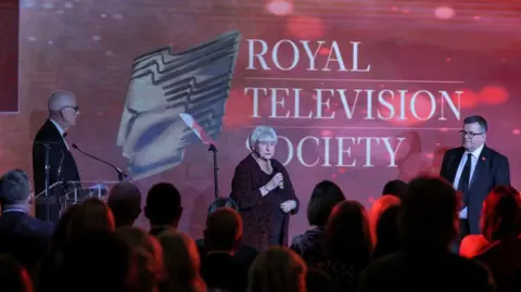 North News and Pictures Ann stands on the stage and speaks into a microphone, while a crowd watches.