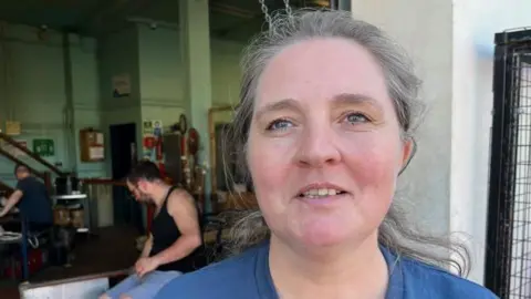 Head and shoulders of a smiling woman with dark, greying hair, wearing a blue t-shirt. In the background is a glassware factory, with workers shaping materials. 