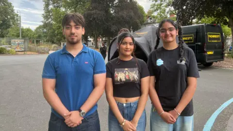 A photo of three young people looking into the camera with small smiles. They have all got their arms crossed Infront of them. There is a boy on the left with a blue top on, and two girls with black tops next to him. 
