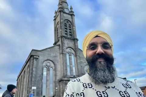 Galab is wearing a yellow turban. He has a beard and glasses. In the background is the stone built gurdwara.