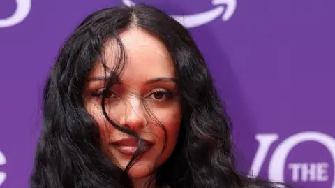 Reuters Jade Thirlwall is standing in front of a purple background with a few strands of her hair in front of her right eye.