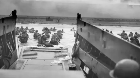 Getty Images A black and white image of the D-Day landings taken from the boat. Servicemen wade through the water with rucksacks and head towards the beach.