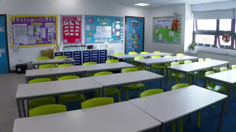 BBC An empty classroom. There are rows of seats which are green in colour. There are signs on the boards around the classrooms of children's work.