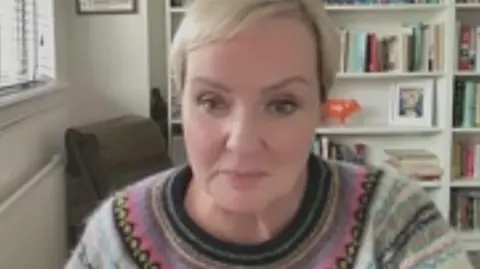 Zoom call from Susan Browne. She is wearing a wool sweater with a Scandi pattern. She has short blonde hair. Behind her there is a white bookshelf with books, decorative items and photos. 