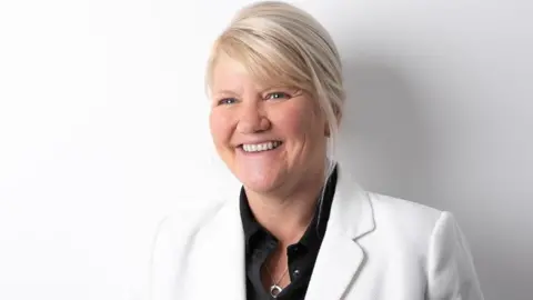 Visit Isle of Man Deborah Heather has fair hair tied back. She's wearing a black shirt with a white suit jacket and is smiling. There's an all white background.