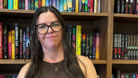BBC/Eve Connor Hayley Beckinsale. She has long, dark brown hair, and is wearing tortoiseshell glasses and a brown vest top. Behind her is a bookcase, and three shelves are visible. Each is full with an array of books in different colours and genres.

