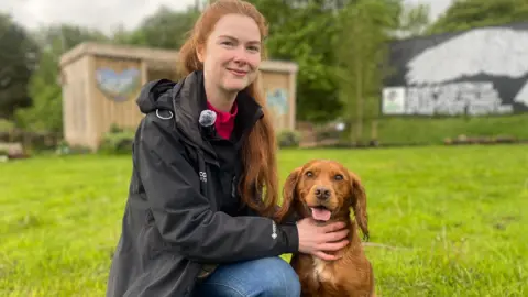 A woman crouches on one knee. She wears a black waterproof coat and a red jumper under it. She wears blue jeans and brown boots as she holds a dog, with both hands, that sits to her left. They pose on green grass with a wooden shed in the background. 