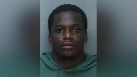 A man wearing a green hooded jumper stares directly at the camera
