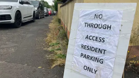 A sign rests against the wall. It stands in front of a number of parked cars and reads: No through access, resident parking only