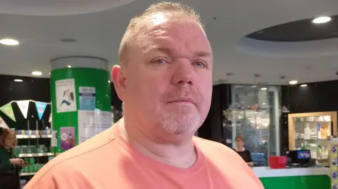 Christopher Connor stands in a pharmacy. He is wearing an orange t-shirt with black writing that says 'silverback'. He has grey hair and blue eyes.