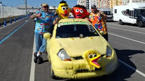 John and Mark Everett A furry yellow car that looks a bit like Big Bird from Sesame Street. There are two men standing next to the car giving a thumbs up. They are both smiling. On top of the car there are Bert and Ernie characters. Behind them are some coaches. They are on the road.