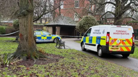 A police car and van in an area of an urban park, with police tape cordoning off a brick shelter