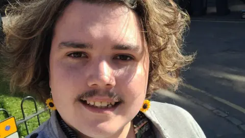 Dylan Lewis-Creser Dylan smiles in a selfie on a sunny day while wearing sunflower earrings 