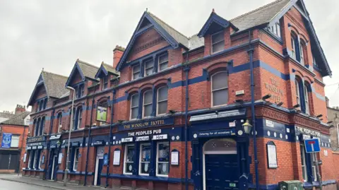 General view of the pub which is a brick building with blue wrap around signs which have the pub's name