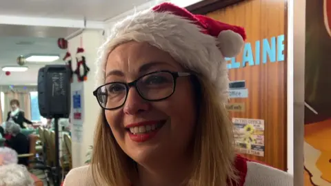 Claire Garner is wearing a red and white Santa hat and is wearing a red and white jumper. She is wearing large, black-rimmed glasses.