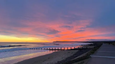 BBC Weather Watchers/Mitchell Small Redcar beach at high tide with the waves reaching most of the way up the sandy beach. The sky is a mixture of blue, orange, yellow and grey with many clouds. A cliff can be seen in the distance and a concrete path to the right.