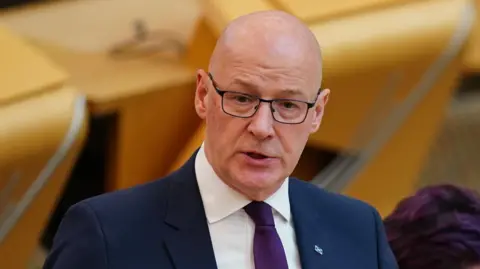 PA Media John Swinney wears dark glasses and wears a navy suit with a white shirt and purple tie in the debating chamber
