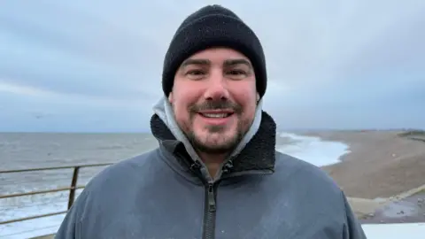 A man smiling wearing a black hat and grey rain coat on the beach. 