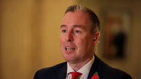PA Media Paul Givan, a man with dark hair, wearing a black jacket, with white shirt and red tie.