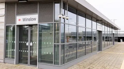 East West Rail Winslow railway station, a grey building with lots of windows, in a box shape, with a paved front.