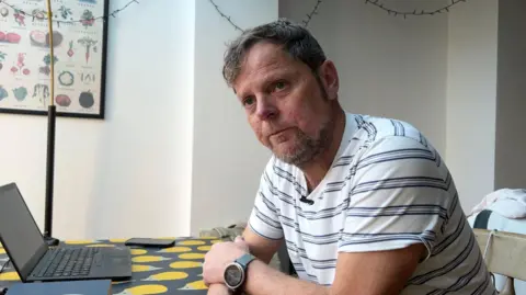 Chris O'Neill. He is sitting at a desk with a laptop in front of him. He has dark hair and a beard. He is wearing a white T-shirt with a blue hooped pattern.