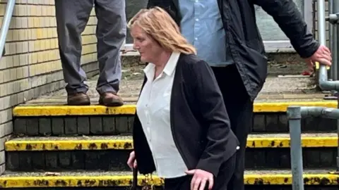 Looking at a woman from a side angle, she is wearing a white shirt and black blazer walking down a set of steps. Two faceless people behind her walking down the steps, holding the handrails. 