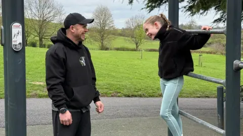 father and daughter James Robinson and Thea Robinson looking at eachother smiling whilst playing on an outdoor gym