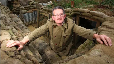BBC A man in a brown, army like coat standing in a trench. He has brown hair and has glasses and a moustache.