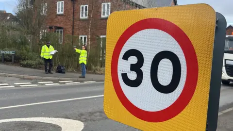 George Carden/BBC Lewes Road with a 30mph zone sign near some houses. Two volunteers are standing on the other side with a speed gun