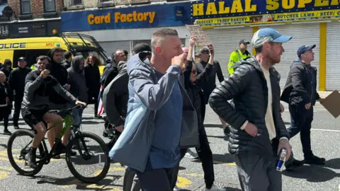 Jonny Humphries/BBC A group of protesters walk down a street. One man is on a pedal bike, apparently filming with his phone, and a woman is carrying a cardboard sign reading 'Liverpool stands with Belfast'.