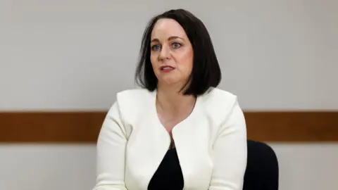 PA Kierans has a black bob and wears a white cardigan and black top. 