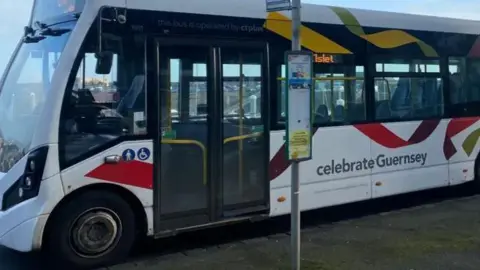 Picture of a bus at a stop in Guernsey. It's a white bus with red, yellow and green swirls.