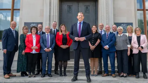 Welsh government The first minister and cabinet ministers stand on steps in front of a grey building with a brown door.