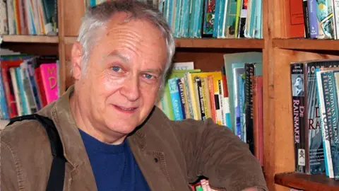 Phil Rickman, who has short grey hair and blue eyes, standing next to a bookcase. He's wearing a brown jacket and blue jumper 