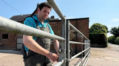 BBC Dave Eardley is dressed in a blue t-shirt under black dungarees. He is locking a silver farm gate with a chain.