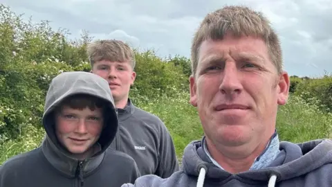 Glen Annison Glen Annison is at the front of the image, taking a selfie. He is a man in his 40s with a weatherworn face and wearing a blue top. His sons stand behind him, the youngest Tyler is wearing a grey top with the hood up and smiling, the oldest Joshua also has a grey top and has fair hair.