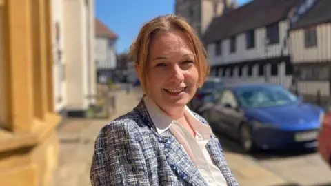 A white woman smiling toward the toward the camera, she is wearing white shirt and blue jacket and standing on street pavement , with cars parked behind her.