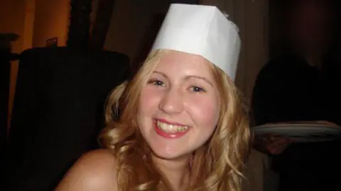 BBC/Alleycats TV A woman smiles at the camera. She wears a white party hat and has long, curly blonde hair.
