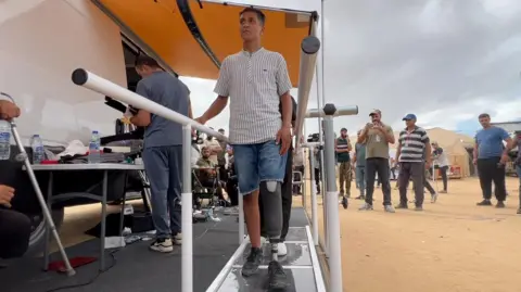 Rizeq Tafish walks using his new prosthetic leg near a special stage with long handles with a crowd of people around him.