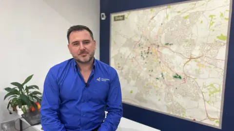 Oliver Ibbotson, the managing director of Derby letting agency Houseshare Heroes, purchases on a table in a blue shirt infront of a map of the city where the new Article Four Direction area is circled 