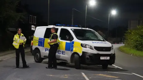 BBC/Oli Constable Two police officers stand in front of a police van blocking the road