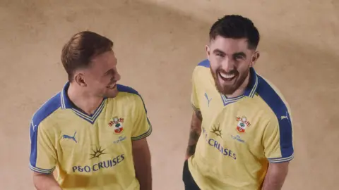 Leo Scienza and Ryan Manning are both wearing the new yellow and blue commemorative shirts. They're both laughing. Leo looks over at Ryan while Ryan grins up towards the camera. The shirts feature a P&O Cruises logo and the words '1976 FA Cup winners'. 