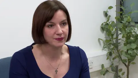 A woman with mid-length dark hair, a silver necklace and a blue top sits on a chair next to a plant.