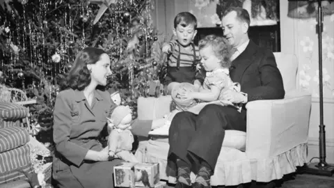 Getty A black and white image of a family sitting around the Christmas tree. The father, a man in a suit with dark hair is on an armchair and has a young girl sitting on his knee. She has short curly blonde hair and is holding a drum. Her mother is sitting on the floor knelt down with a doll on her knee. Their son, a boy of about three, is sitting on the arm of the chair next to his father.