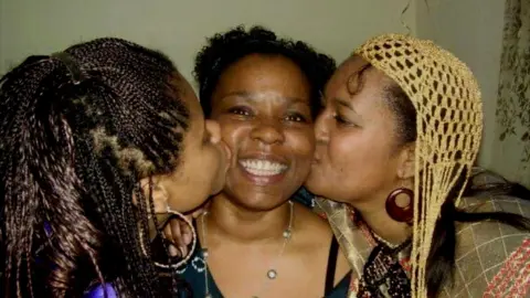 three women, two of them are kissing the third on the cheek