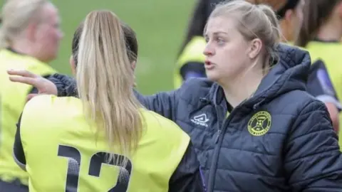 Adam Reapa Laura Broadbent wears a puffa-style jacket, bearing the Leafield Athletic logo - a yellow circle surrounding a football. Laura looks like she is giving some stern advice to a player, whose back is to the camera. The player wears a bright yellow bib with the number 13 partially visible on the back.