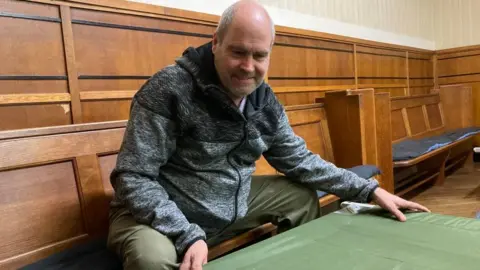 Martin Heath/BBC Man wearing a grey sweater setting up a campbed